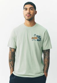 Vihreä puuvillapaita, jossa on graafinen kuvio, jossa lukee "Ride the Wave" ja surffaaja, lyhyet hihat, pyöreä kaula-aukko, sileä pinta.