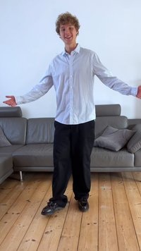 Camicia azzurro chiaro a righe bianche con bottoni, pantaloni neri e scarpe nere lucide. In piedi su un pavimento di legno in un interno moderno.