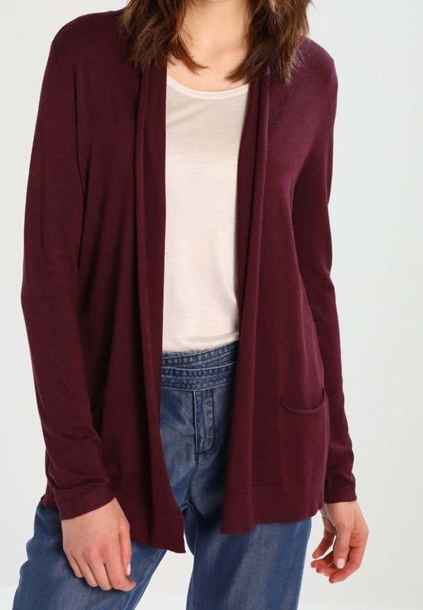 Femme portant un cardigan bordeaux ouvert sur un haut blanc et un jean taille haute bleu, debout devant un fond uni.