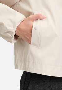 Jasnobeżowa kurtka ze ściągaczem na rękawie, boczną kieszenią na zatrzask i gładką teksturą, połączona z ciemnym, wzorzystym dołem.