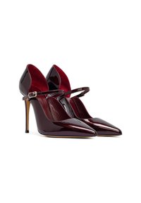 Un par de tacones altos de charol rojo oscuro con punta afilada y delgadas correas en el tobillo contra un fondo blanco.