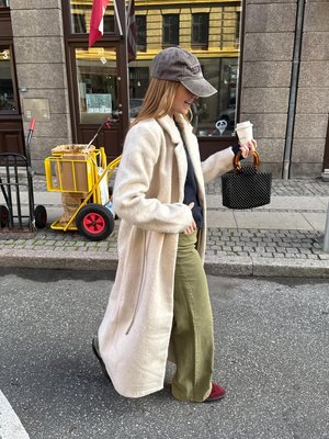 Moteris dėvi ilgą, kreminės spalvos, tekstūruotą paltą, alyvuogių žalių plačių kelnių ir bordo spalvos batus, rankoje laiko juodą tinklelio rankinę ir kavą.