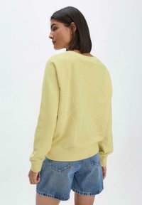 Gelbes Sweatshirt mit lockerer Passform, auf der Rückseite mit weißer Blumenstickerei versehen, kombiniert mit Jeansshorts. Weicher Stoff und gerippte Bündchen.