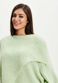 Light green knit poncho with a textured finish, featuring a loose, draped design and a round neckline.