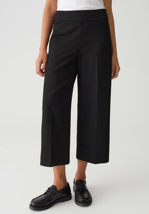 Pantaloni cropped neri a gamba larga con una texture liscia, caratterizzati da una vita alta. Abbinati a scarpe nere con laccio.