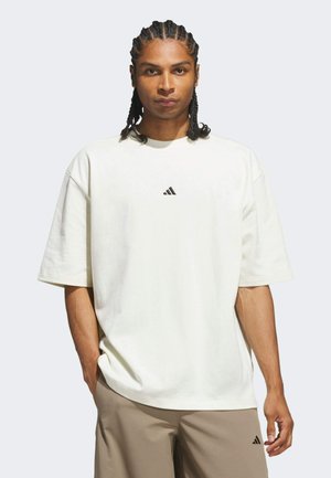 Giovane uomo con capelli intrecciati che indossa una maglietta bianca larga Adidas e pantaloncini beige Adidas, in piedi con una mano in tasca.