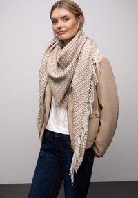 Jasnobeżowy pleciony szalik z teksturowanym wzorem i frędzlami na krawędziach, owinięty wokół szyi, w połączeniu z beżową kurtką i ciemnymi dżinsami.