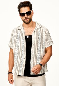 Kurzarmhemd in Creme mit braunen vertikalen Streifen, strukturiertem Stoff und schwarzen Knöpfen. Getragen über einem schwarzen Tanktop, kombiniert mit Sonnenbrille.