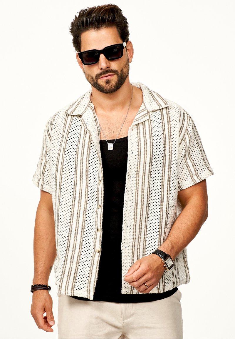 Kurzarmhemd in Creme mit braunen vertikalen Streifen, strukturiertem Stoff und schwarzen Knöpfen. Getragen über einem schwarzen Tanktop, kombiniert mit Sonnenbrille.