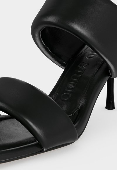 Μαύρο δερμάτινο πέδιλο με ψηλό τακούνι, δύο επενδεδυμένους ιμάντες και λεπτό τακούνι σε λευκό φόντο.