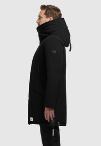 Sort lang parka med høj krave og hætte. Har en frontlynlås, sidelommer og brandede detaljer. Glat, mat stofstruktur.