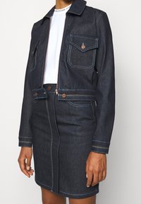 Mörk denimjacka med två bröstfickor, koppardetaljer och blå sömmar. Bärs över en vit topp, matchat med en denimkjol.