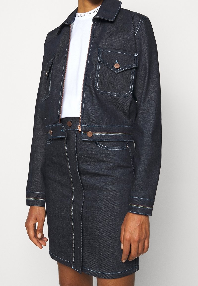 Mörk denimjacka med två bröstfickor, koppardetaljer och blå sömmar. Bärs över en vit topp, matchat med en denimkjol.