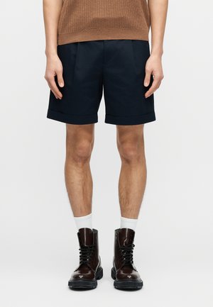 Shorts bleu marine à texture lisse, avec un ourlet replié et des plis. Associés à des bottes en cuir marron et des chaussettes blanches.