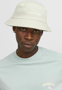 White bucket hat with "Originals Studio" logo, made of fabric with a smooth texture. Worn with a light blue t-shirt.