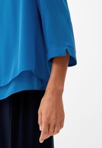 Bras droit et main d'une personne portant un chemisier bleu vif à manches longues avec une petite fente latérale au niveau du poignet et un pantalon foncé.