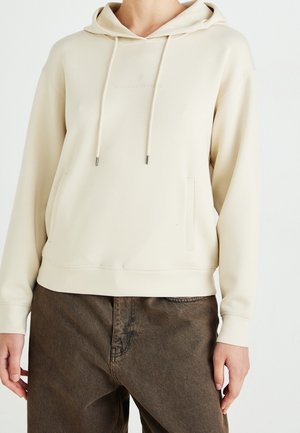 Personne portant un sweat à capuche beige avec des poches avant et un pantalon marron sur un fond blanc uni.