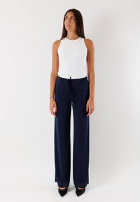 Top bianco senza maniche e pantaloni larghi blu navy con vita regolabile. Il modello indossa scarpe nere a punta su uno sfondo uniforme.