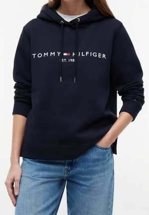Personne portant un sweat à capuche Tommy Hilfiger bleu marine avec un logo texte blanc et rouge et un jean bleu clair, une main dans la poche.