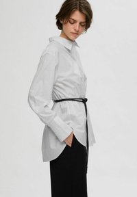 Camicia a righe bianche con una cintura nera in vita, maniche lunghe e colletto a punta, abbinata a pantaloni neri.