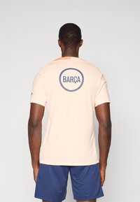 Licht perzik sportshirt met een ronde "BARÇA" logo in marineblauw, korte mouwen en een ronde halslijn. Draagt bij navy shorts.