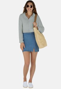 Hellgrünes Hemd mit Knöpfen, blauer Denim-Rock mit Schlitz, weiße Slipper, runde Sonnenbrille und eine geflochtene Strohtasche.