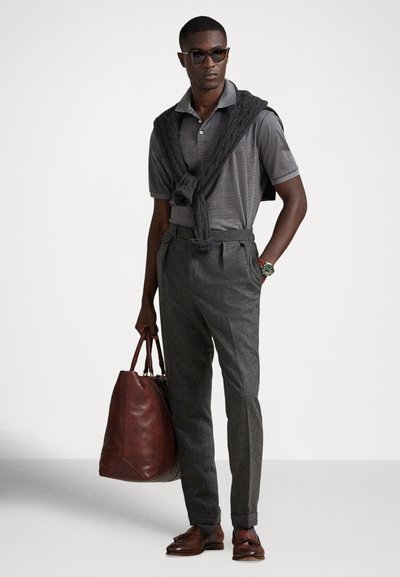 Άνδρας που φορά γκρι πόλο, σκούρο γκρι πουλόβερ περασμένο πάνω από τους ώμους, γκρι παντελόνια, καφέ δερμάτινα παπούτσια και τσάντα, και γυαλιά ηλίου, στέκεται με τα χέρια στις τσέπες.