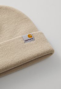 Beige knit beanie with a folded cuff and a rectangular gray label featuring a yellow logo and red text. Soft texture and snug fit.