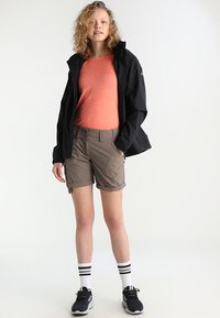 Zwarte waterdichte jas, oranje getextureerde shirt, beige shorts met opgerolde manchet, zwarte sportschoenen en witte sokken met zwarte strepen.