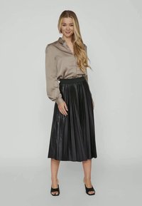 Blusa de cetim bege claro com colarinho e mangas bufantes, combinada com uma saia midi plissada em imitação de pele preta. Sandálias abertas pretas.