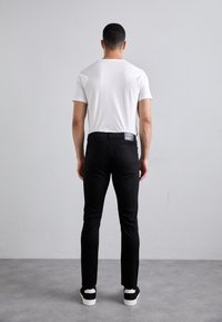 Černé slim-fit džíny s hladkou texturou, ve spojení s bílým tričkem s krátkým rukávem a černými teniskami. Obsahují štítek na zadní kapse.