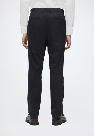 Homme portant un pantalon noir sur mesure, une chemise blanche à manches longues et des chaussures habillées marron foncé, vu de dos sur un fond uni.