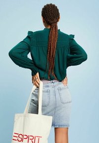 Femme aux longs cheveux tressés, portant un chemisier vert et une jupe en jean, tenant un sac fourre-tout blanc avec le logo rouge « ESPRIT » dans son dos.