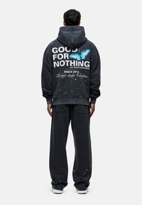 Zwarte hoodie met de tekst "GOOD FOR NOTHING" en een vlinderafbeelding op de achterkant; gecombineerd met bijpassende zwarte joggingbroek met verfspatten.