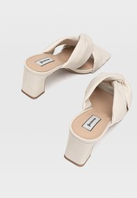 Mules à talons beiges avec un dessus en tissu torsadé et un bout carré. Présente un talon bloc épais et une semelle intérieure rembourrée pour le confort.