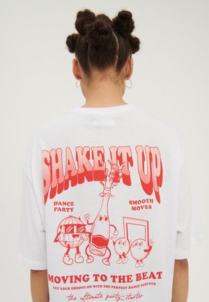 Osoba z warkoczami, ubrana w białą koszulę z czerwonymi postaciami z kreskówek oraz napisami "Shake It Up" i "Moving to the Beat" na plecach.