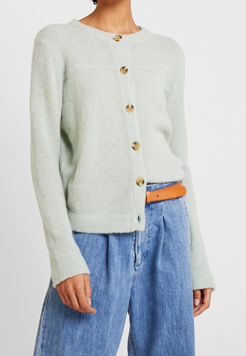 Cardigan en maille vert clair à col rond, avec cinq boutons beiges sur le devant et des poignets côtelés, associé à un pantalon en denim bleu.