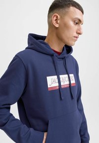 Marineblå hoodie med en frontkangaroo lomme, justerbar snorhætte og et grafisk logo patch i hvid og bordeaux. Blød tekstur.