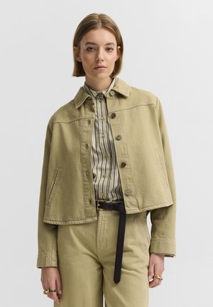 Frau mit kurzen Haaren, die eine beige kurze Jacke, ein gestreiftes Hemd mit Knöpfen, beige Hosen, einen schwarzen Gürtel und Ringe an den Fingern vor einem einfarbigen Hintergrund trägt.