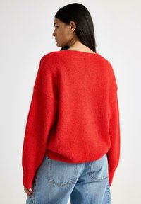 Červený nadměrný svetr z měkkého, strukturovaného pletení, s širokým výstřihem a žebrovanými manžetami, doplněný světle modrými džínami s vysokým pasem.