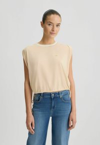 Top beige a maniche corte con orlo arricciato, dotato di un colletto bianco. Tessuto morbido con una leggera lucentezza, abbinato a jeans in denim blu.