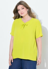 Blusa verde lime a maniche corte con scollo a V e dettaglio fiocco sul davanti; tessuto leggero e morbido; spacchi laterali per facilitare i movimenti.