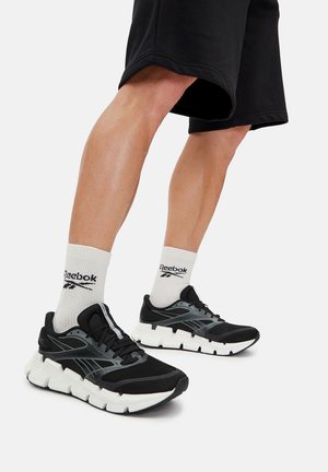 Czarne buty do biegania z teksturowaną siateczkową cholewką; jasnoszare akcenty; gruba biała podeszwa; zestawione z białymi skarpetkami Reebok.