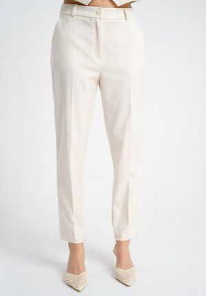 Kobieta w wysokich, dopasowanych kremowych spodniach i beżowych plecionych szpilkach z zaostrzonym noskiem, stojąca na tle jednolitego białego tła.