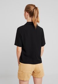 Donna con coda di cavallo bionda, che indossa una maglietta nera a maniche corte e pantaloni corti beige, in piedi di spalle contro uno sfondo grigio chiaro uniforme.