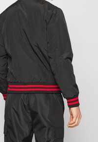 Svart bomberjacka med rödrandiga ribbade ärmslut och midja. Slät, glänsande tyg. Har en strukturerad, lätt design.