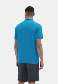 Polo shirt blu a maniche corte con un design rigoroso, texture liscia e dettagli gialli a contrasto nel colletto. Indossato con pantaloni corti scuri.