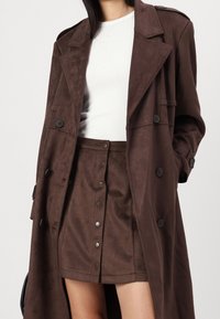 Kvinna iklädd en brun knapp-frontsuede kjol, matchande lång ytterrock och vit ribbad långärmad topp, stående med en hand i ytterrockens ficka.