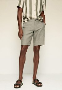 Uomo che indossa pantaloni corti grigi chiari, una camicia a righe verticali verde e bianca a maniche corte e sandali neri con doppia fibbia, con le dita dei piedi visibili.