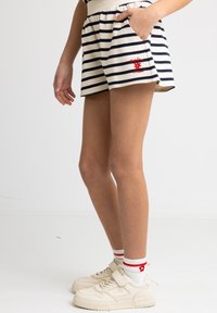 Spodenki w paski w kolorze kremowym i granatowym z elastycznym paskiem w talii, bocznymi kieszeniami oraz czerwonym haftowanym logo na lewej nodze. Połączone z kremowymi trampkami.